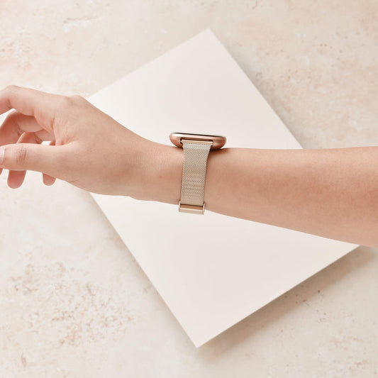 Gold Slim Milanese Coconut Lane Fitbit strap on a wrist, beige background, white card beneath the hand.