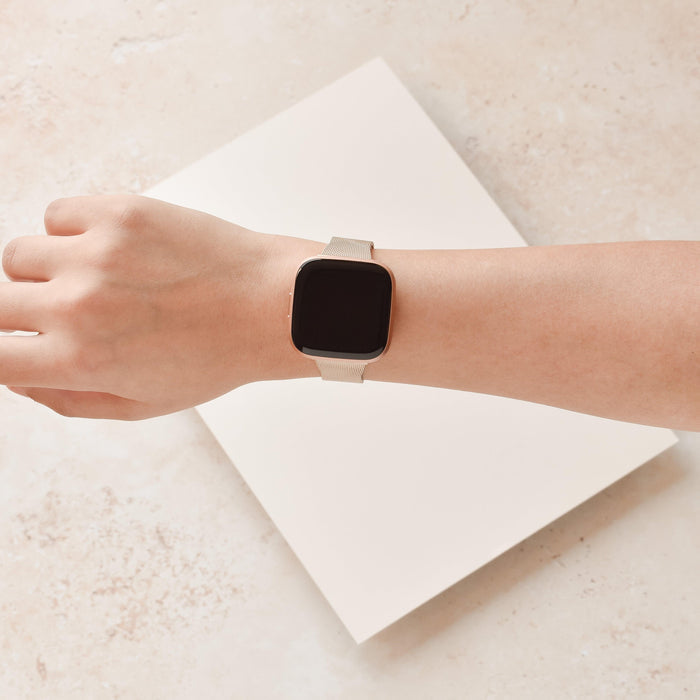 Gold Milanese Fitbit strap on a square-faced smartwatch, worn on a left hand against a beige background.
