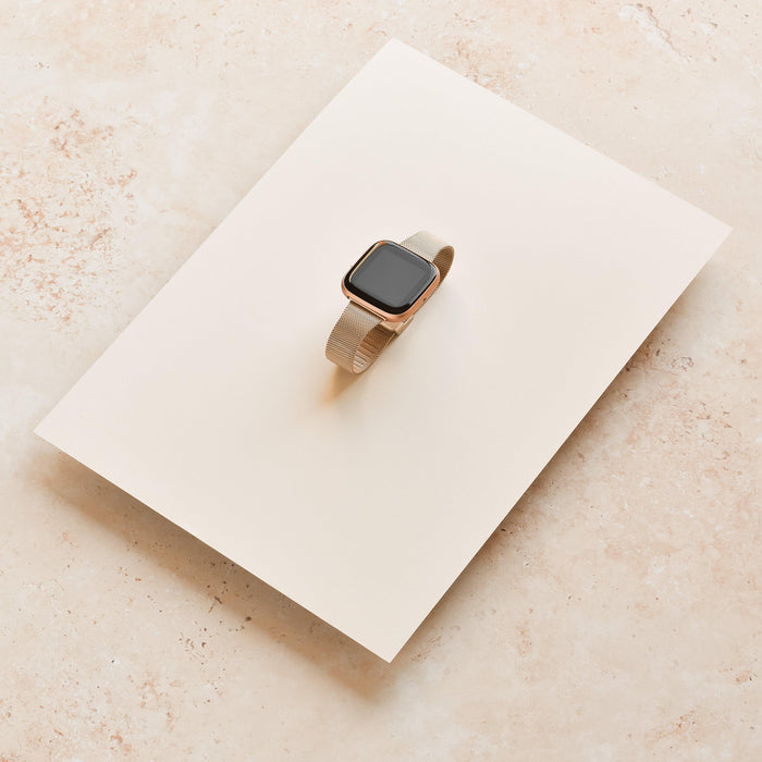 Beige smartwatch with blank screen and gold Slim Milanese metal strap, placed on a cream-colored sheet.
