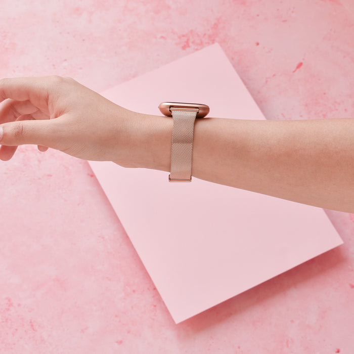 Hand wearing a slim rose gold milanese Coconut Lane Fitbit strap against a pink background.