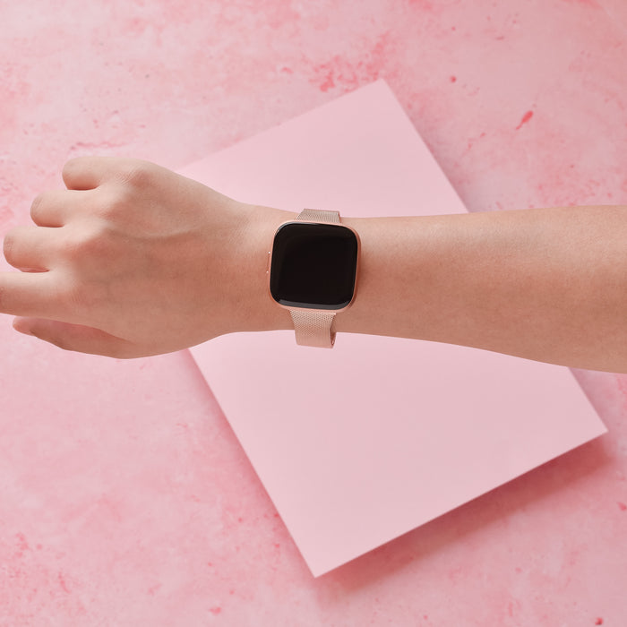 Hand wearing a rose gold Slim Milanese Fitbit strap, shown on a pink surface with pink paper in the background.