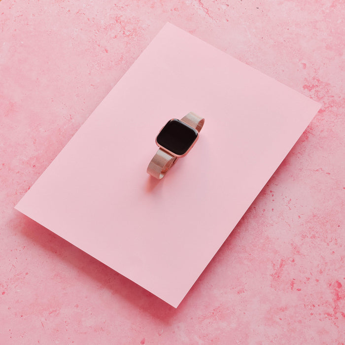 Rose gold slim Milanese Coconut Lane Fitbit strap displayed on a pink sheet with a textured pink background.