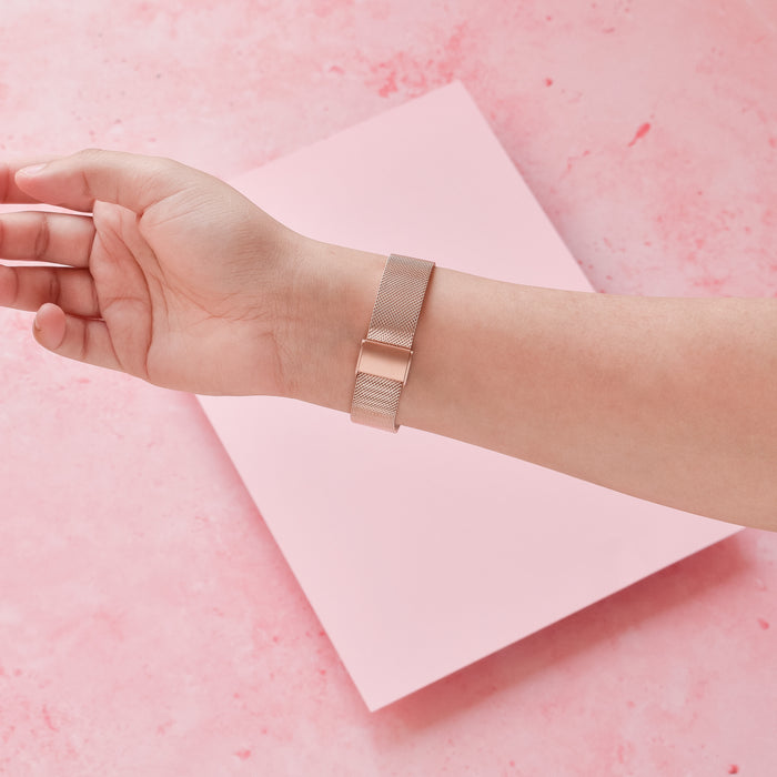 Slim rose gold Milanese Coconut Lane Fitbit strap on a hand, against a pink background with pink paper accents.