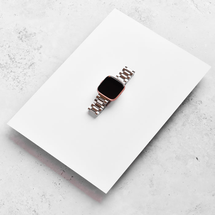 Silver stainless steel Coconut Lane Fitbit strap displayed on a white sheet over a light gray textured background.