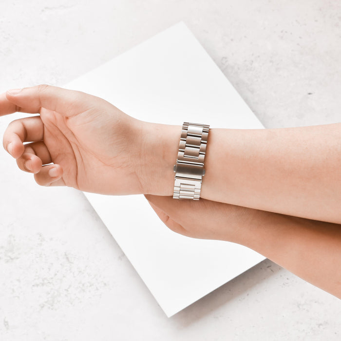 Silver stainless steel Coconut Lane Fitbit strap worn on a hand, displayed against a white background.