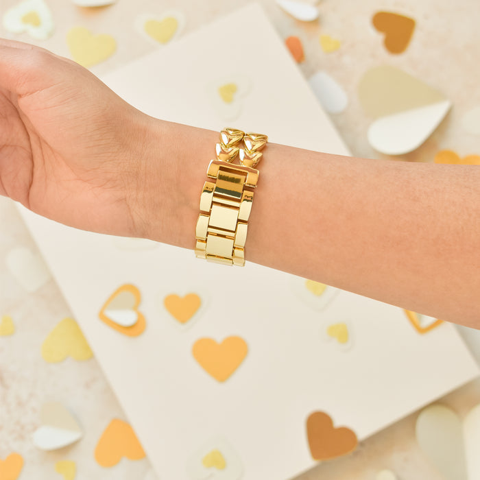 A hand adorned with Coconut Lane's Hearts Chain Apple Watch Strap - Gold is displayed against a backdrop sprinkled with gold, white, and beige heart-shaped confetti. The off-white surface in the scene adds a soft and elegant touch to the presentation.