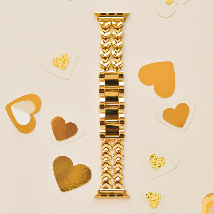 An intricately designed gold watch band is laid flat on a light-colored surface. Surrounding the Coconut Lane Hearts Chain Apple Watch Strap - Gold are assorted heart-shaped decorations in gold and white, with some featuring glitter accents. This elegant stainless steel adjustable strap fits various Apple Watch models.
