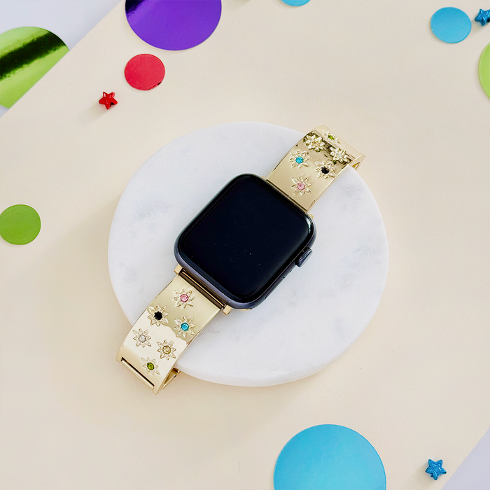 Gold Apple Watch strap with colorful stone links and floral designs, displayed on a background of vibrant confetti.