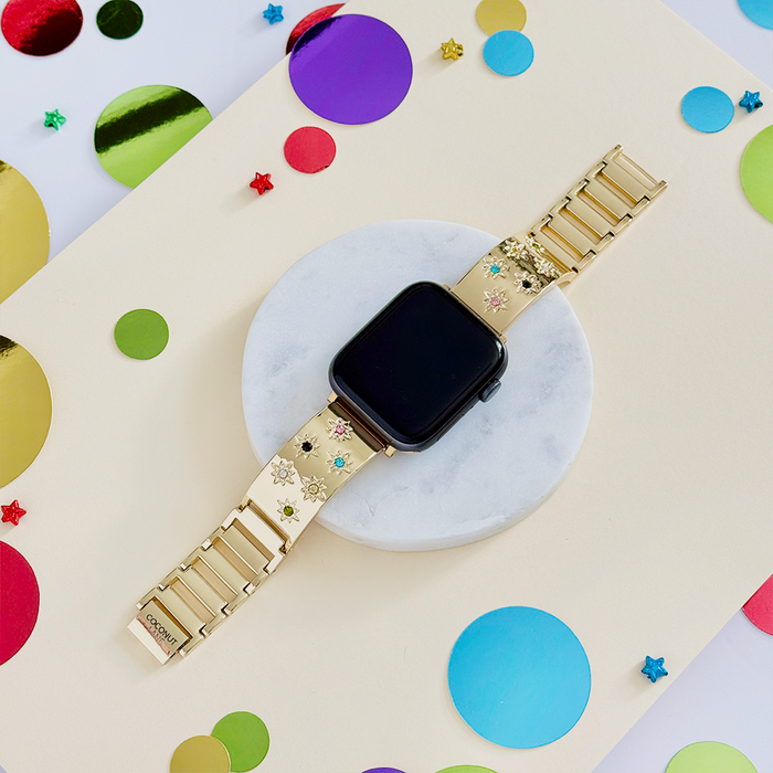Gold and coloured stone Apple Watch strap displayed on a round white platform with scattered confetti.