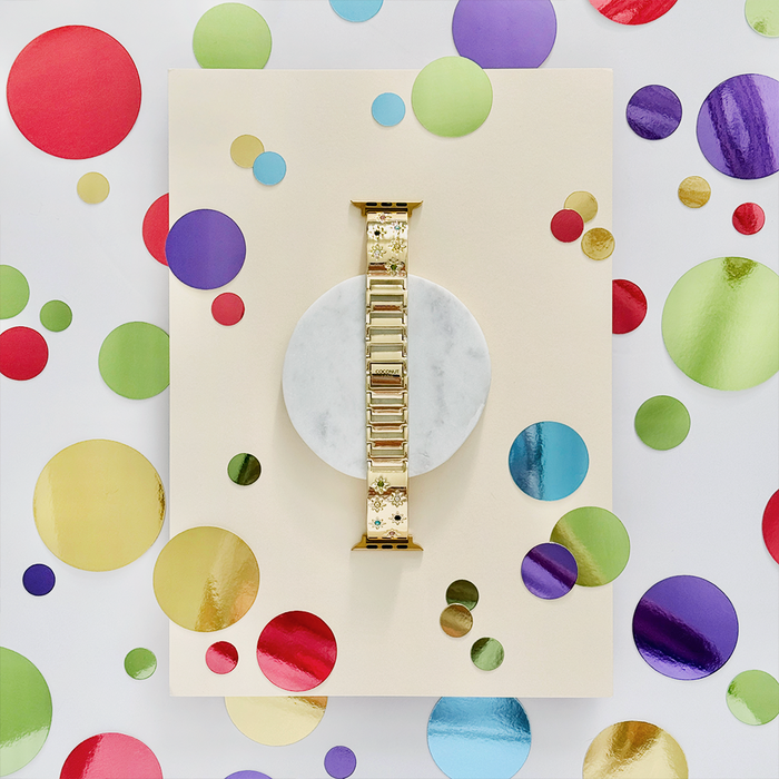 Gold Apple Watch strap with colourful stone accents on marble, surrounded by metallic confetti circles.
