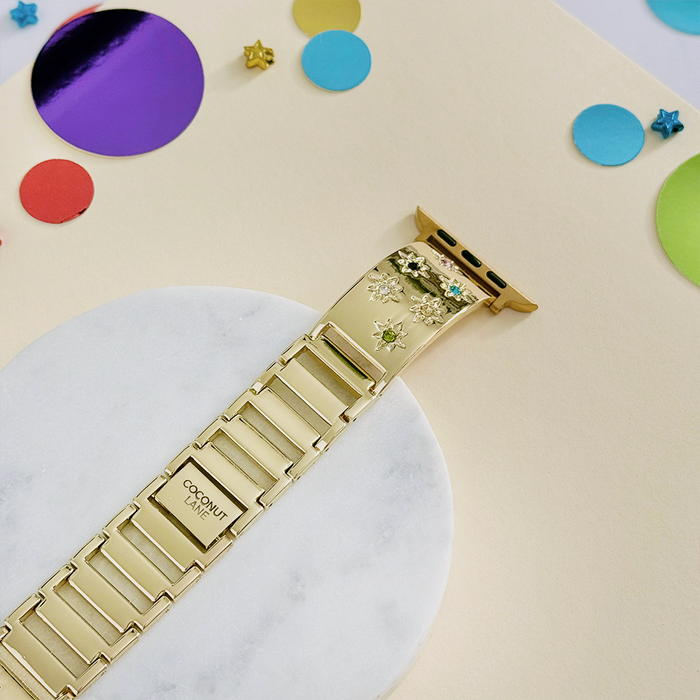 Gold Apple Watch strap with multicoloured stone links, displayed on a marble surface with bright paper circles.