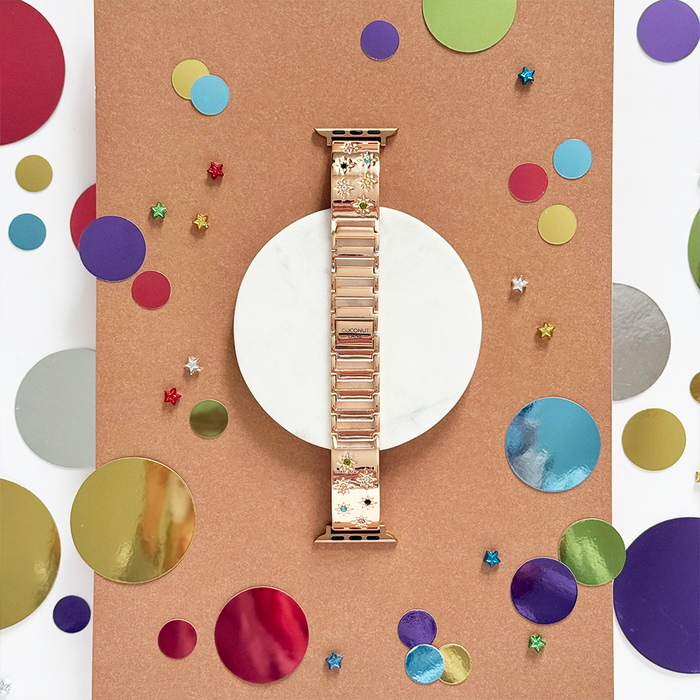 Rose gold Apple Watch strap with coloured stones, displayed on a white circle with multicoloured dots.