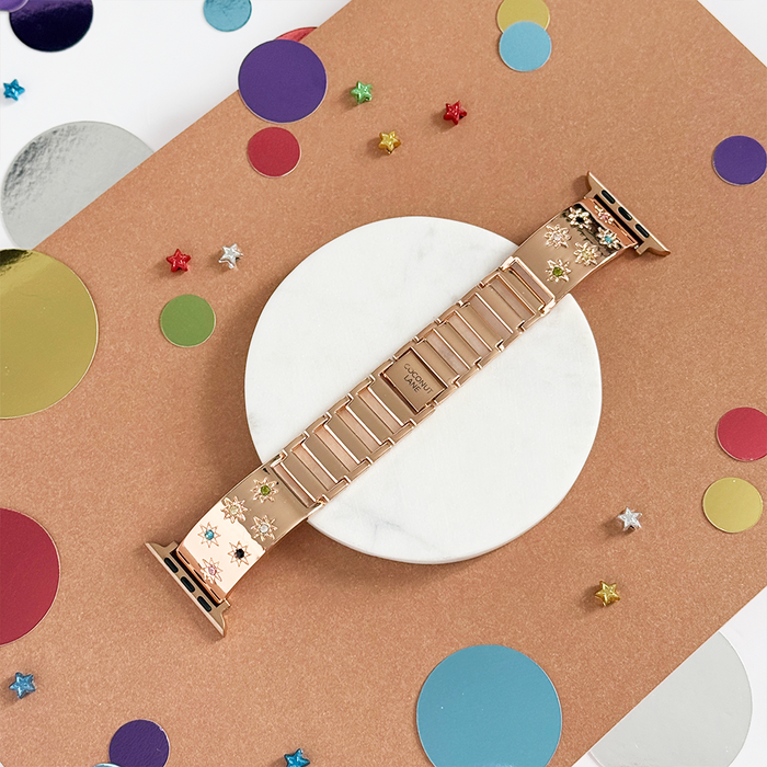 Rose gold Apple Watch strap with coloured stones, displayed on marble with confetti and star accents.