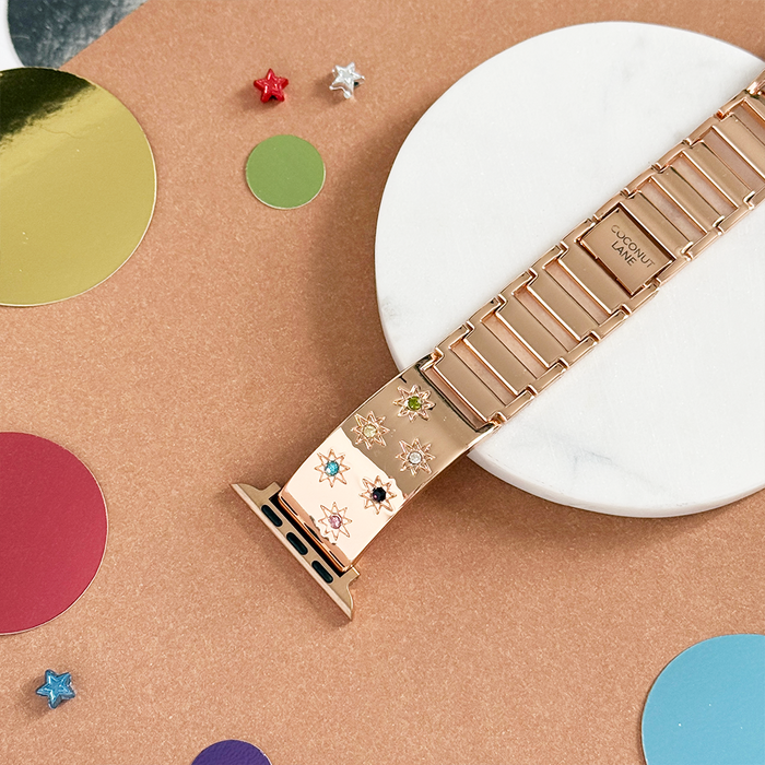 Rose gold Apple Watch strap with colorful stone charms, displayed on a brown background.