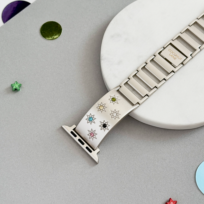 Starlight Apple Watch strap with colored stone links, displayed on marble and gray surface with scattered sequins.