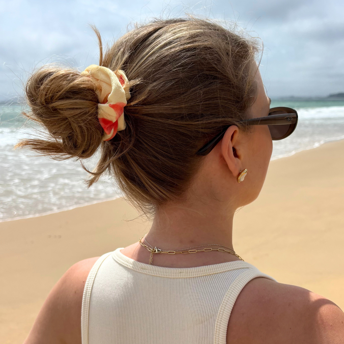 Woman with messy bun wearing a white floral scrunchie stands on a cloudy beach, facing the ocean.