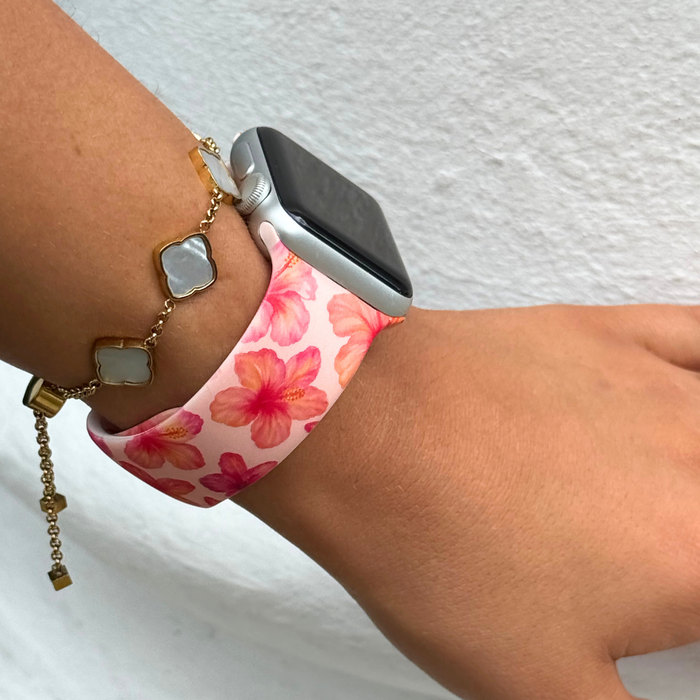 Pink hibiscus-patterned Apple Watch strap on a wrist, paired with a gold bracelet featuring white stones.