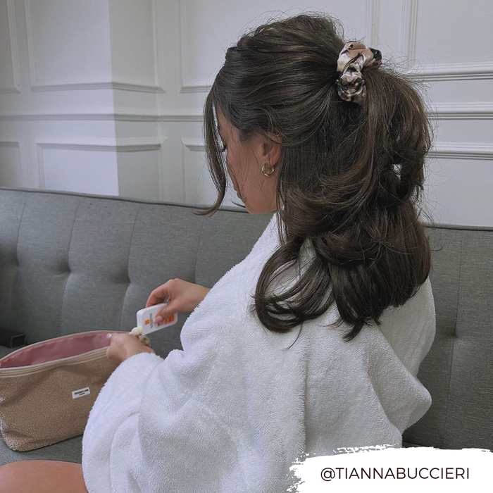 Woman in a white robe on a couch, wearing an ivory tortoiseshell scrunchie and holding a pink pouch.