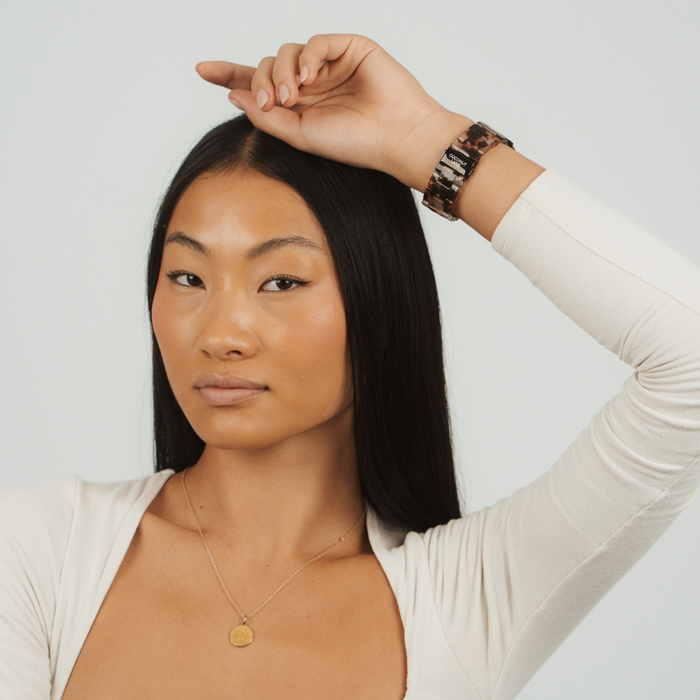 Woman with long black hair wearing a cream top and gold necklace, showing an ivory tortoiseshell Apple Watch strap.