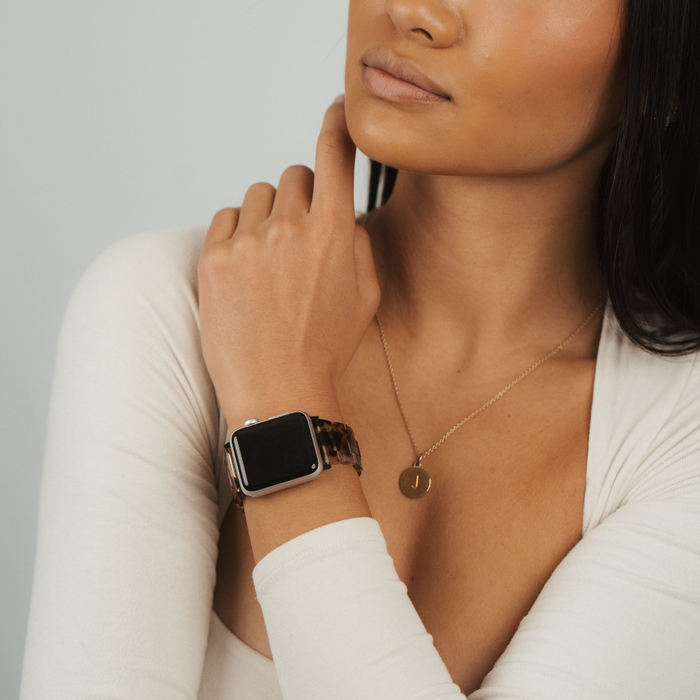 Gold J pendant and ivory tortoiseshell Apple Watch strap on woman in cream top, hand at neck.