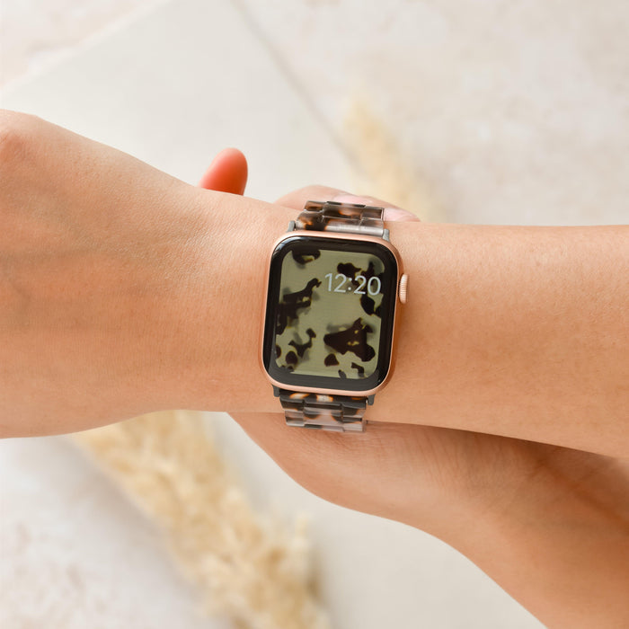 A close-up of a wrist wearing a Coconut Lane smartwatch with the Luxe Ivory Tort Apple Watch Strap and a camouflage-patterned display screen. The time on the watch reads 12:20, and the background is a light-colored, blurred surface.