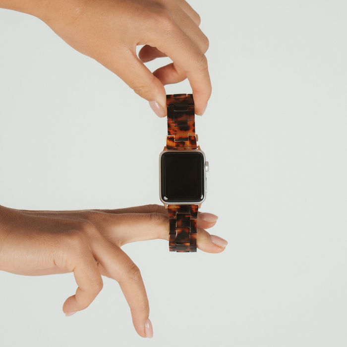 Two hands display a Luxe Tortoiseshell Apple Watch strap with a glossy brown and black pattern against a white background.