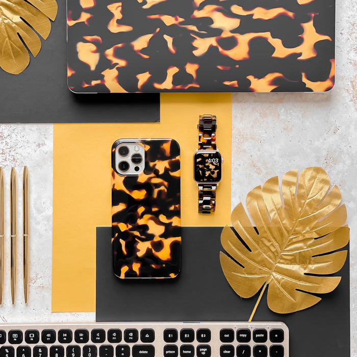 A flat lay features a smartphone, a smartwatch adorned with the Luxe Tortoiseshell Apple Watch Strap by Coconut Lane, and a laptop showcasing matching orange and black swirled patterns. Arranged on a yellow and grey surface, the setup includes gold pens, a black keyboard, and gold-painted tropical leaves for an added aesthetic touch.