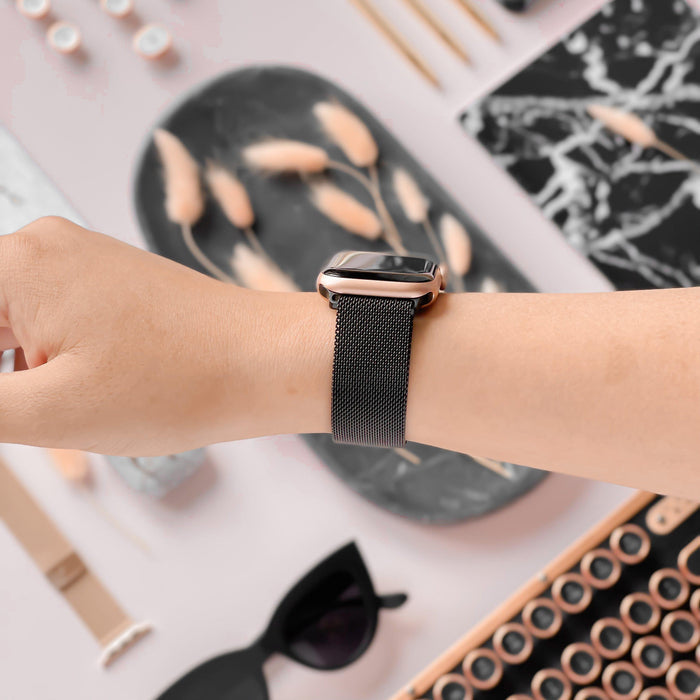 A person’s wrist adorned with the Magnetic Chain Apple Watch Strap in Black by Coconut Lane, featuring a sleek metallic mesh band and a stainless steel clasp. The background showcases an array of stylish items, including sunglasses, a black marble-patterned notepad, and decorative objects tastefully arranged on a light surface.