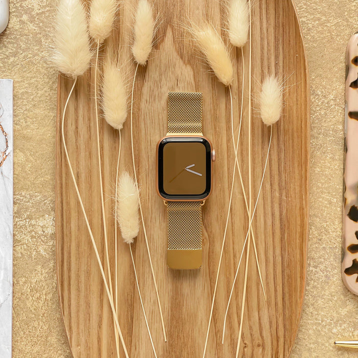 A gold Coconut Lane Magnetic Chain Apple Watch Strap is placed on an oval wooden tray, surrounded by dried beige fluffy grasses. The background features a soft golden-textured surface and parts of other items, including a marble-patterned object and a tortoiseshell object.