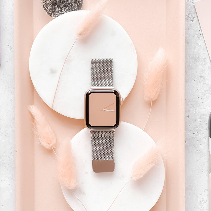 The Coconut Lane Magnetic Chain Apple Watch Strap in Silver rests stylishly across two round marble trays. The background showcases delicate, peach-colored feathery plants, creating a soft and elegant aesthetic. The watch face displays a minimalistic peach-colored design, compatible with various Apple Watch models.