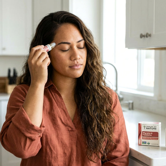 Woman applying a migraine relief roller to her temple in a bright kitchen, product box visible on the countertop