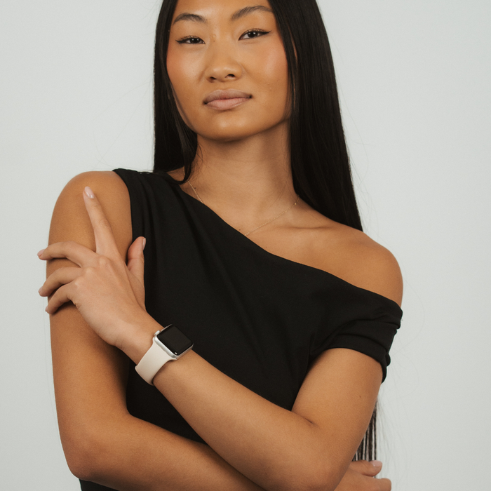 Woman with long black hair wears a black one-shoulder top and an almond white Apple Watch strap.