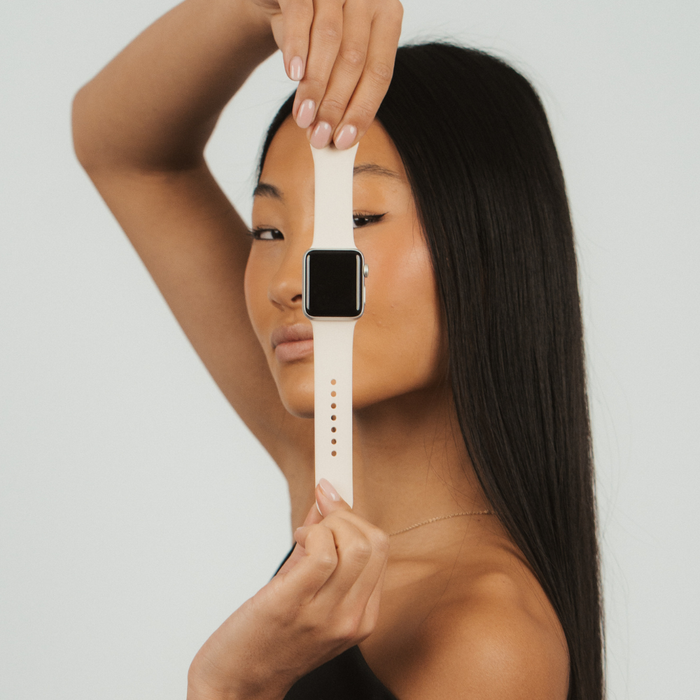 Woman holds an Almond White Coconut Lane NAKD Apple Watch strap vertically, plain background.