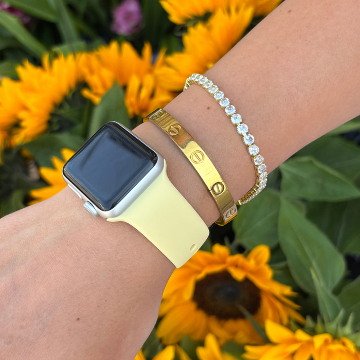 Butter yellow Coconut Lane NAKD Apple Watch strap on wrist with gold bracelets; sunflowers in background.