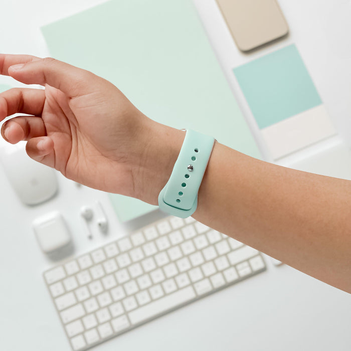 A person's wrist wearing a Cool Mint silicone strap from Coconut Lane's NAKD Apple Watch Strap collection, with a minimalistic workspace background including a white keyboard, mouse, earphones, smartphone, and various stationery items, all in pastel colors.