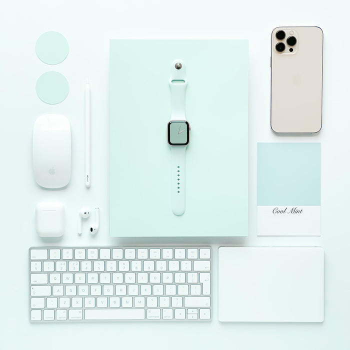 A flat lay of tech gadgets and accessories on a light mint background features an iPhone, Apple Watch with a sleek NAKD Apple Watch Strap in Cool Mint by Coconut Lane, iPad, keyboard, mouse, AirPods, stylus, charger, and a mint-colored card labeled "Cool Mint." The devices are neatly arranged in a grid pattern.