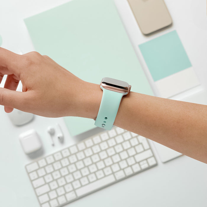 A person's wrist is adorned with a NAKD Apple Watch Strap - Cool Mint by Coconut Lane. The background features minimalistic, color-coordinated items like a keyboard, earphones, and various flat objects in shades of mint green and beige. The overall aesthetic is clean and modern, fitting perfectly with the NAKD Collection.