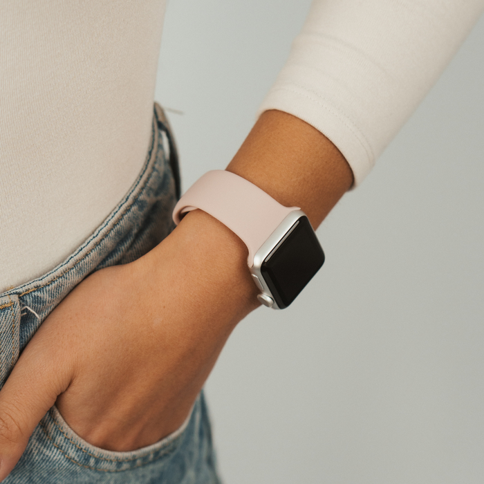 Powder pink Apple Watch strap on a wrist, paired with a white top and blue jeans with one hand in the pocket.