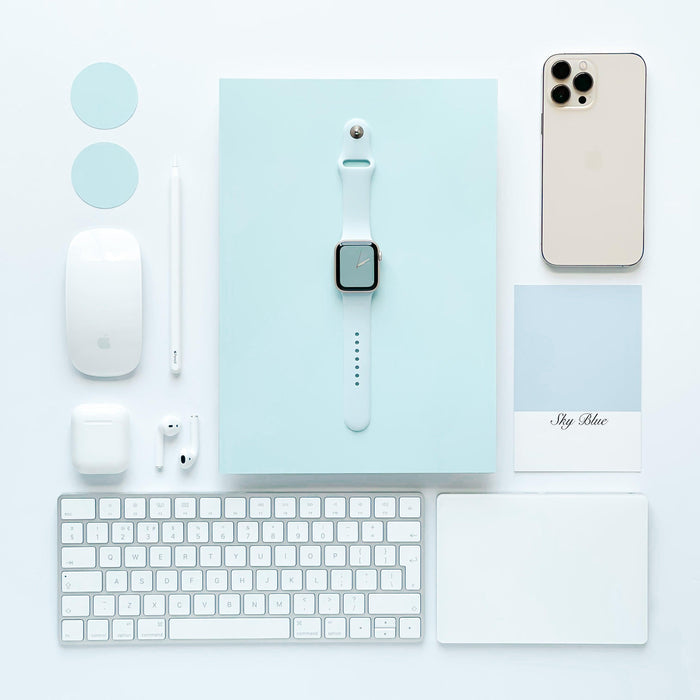A neatly arranged flat lay featuring tech accessories in a minimalistic design. Includes an Apple Watch with a Coconut Lane NAKD Apple Watch Strap - Sky Blue, iPhone, keyboard, mouse, stylus, AirPods, two circular discs, and a card reading "Sky Blue," all set on a light blue rectangular base reflecting spring-inspired pastels.