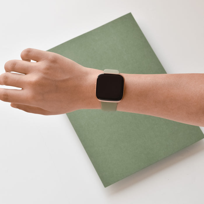 Coconut Lane NAKD Fitbit Strap in Vintage Khaki worn on a wrist next to a green notebook on a white surface.