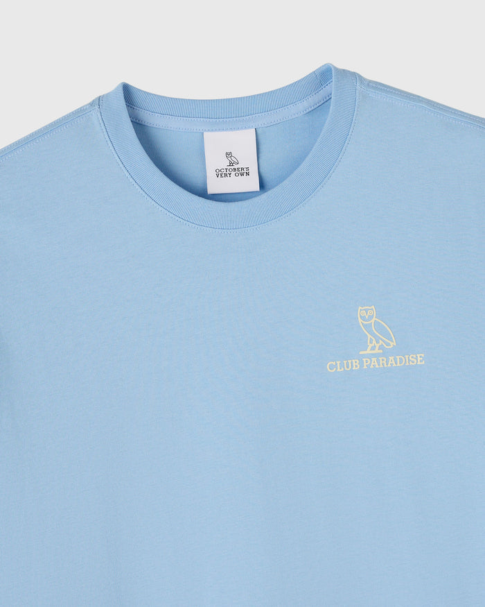 Light blue crewneck tee with OVO owl logo and "Club Paradise" text embroidered on chest.