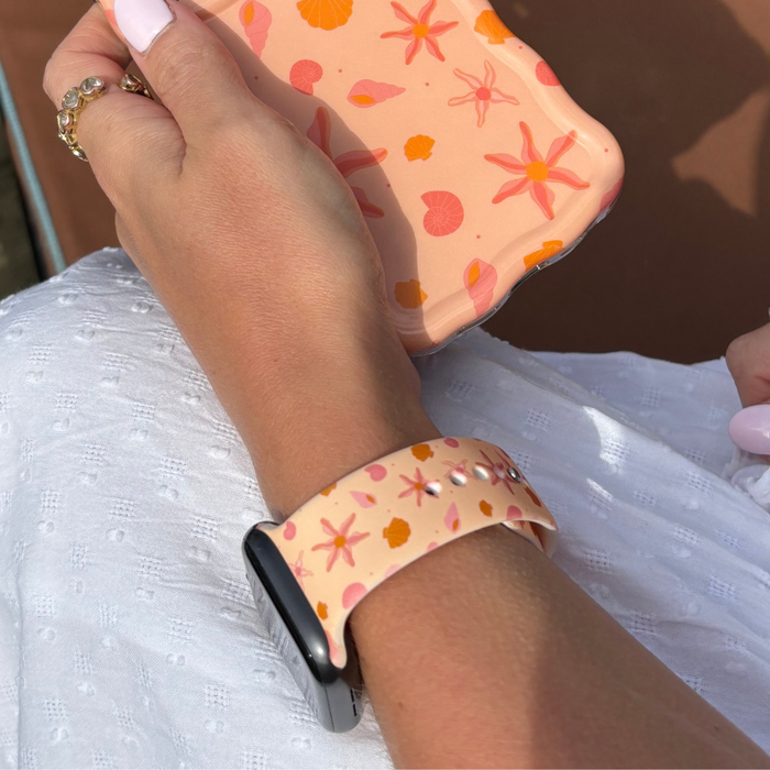 Hand with light pink nails holding a phone, wearing a peach and white Coconut Lane Peachy Beach Apple Watch strap.