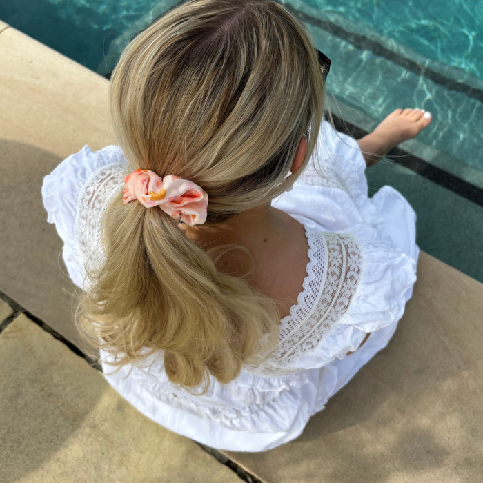 Woman in a white dress sits poolside with a peach and pink scrunchie in her hair, feet near the water.