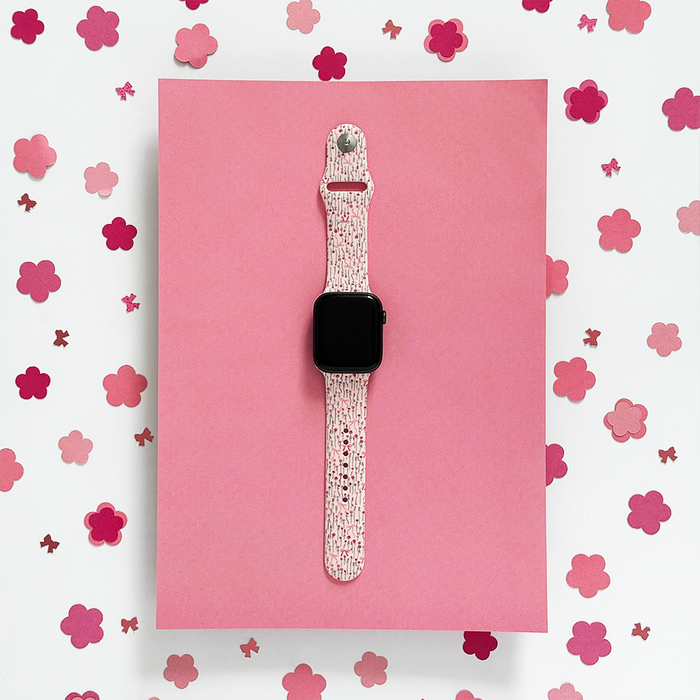 Coconut Lane Peony Bow Apple Watch strap in pink, shown on a pink surface with pink flower cutouts in the background.