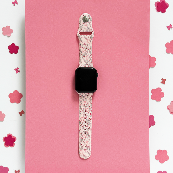 Apple Watch strap with a pink peony bow pattern, shown on a pink surface with scattered flower-shaped confetti.