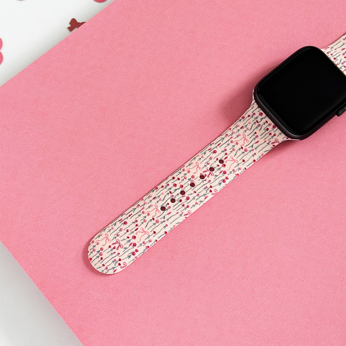Coconut Lane Apple Watch strap with a pink peony bow design on a pink surface, patterned paper in the background.