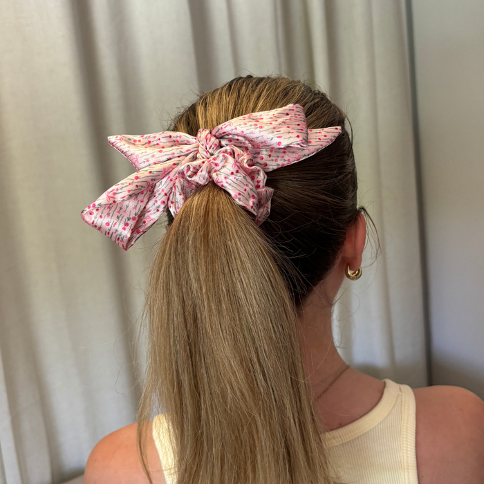 Woman with long blonde hair wears a cream tank and a pink satin bow scrunchie in her ponytail.