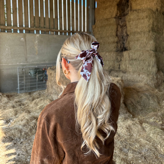 Woman with long blonde hair wears a satin bow scrunchie in a cow print with a brown jacket inside a barn.