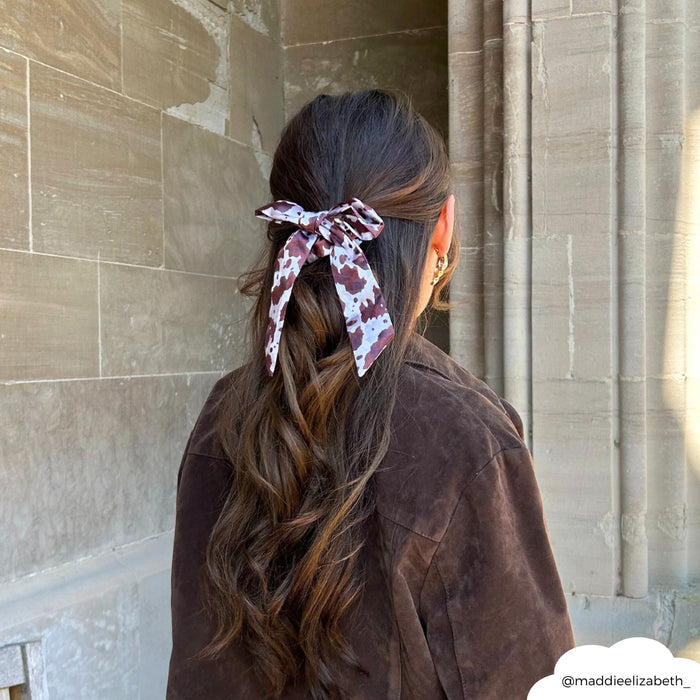 Woman with long wavy hair in a brown jacket wears a brown satin bow scrunchie, standing by a stone wall.