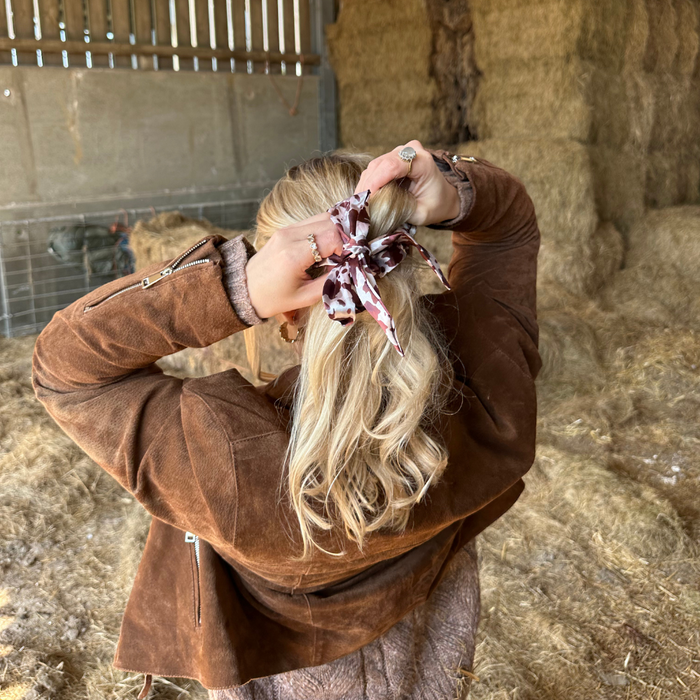 Blonde woman in a brown suede jacket ties her hair with a satin bow scrunchie near hay bales.